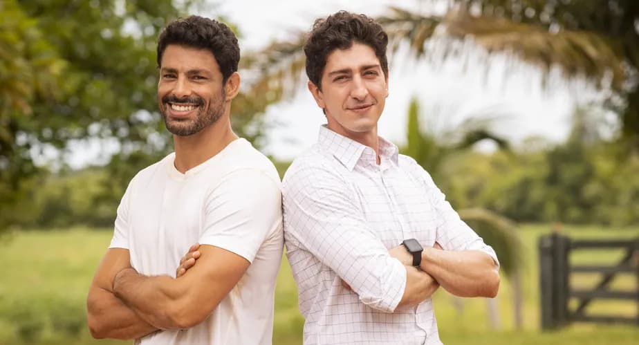 Caio e Daniel, virados de costas um para o outro, vestindo camisa branca sobre um fundo de fazenda na novela Terra e Paixão