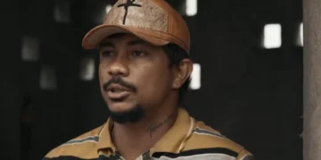 Damião, personagem de Xamã, usando um boné com uma cruz e olhando para a esquerda, em cena de Renascer.