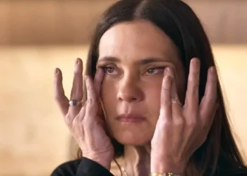 A imagem mostra Mércia, personagem de Adriana Esteves em Mania de Você, com expressão séria e mãos no rosto, pressionando a pele ao redor dos olhos.
