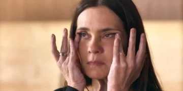 A imagem mostra Mércia, personagem de Adriana Esteves em Mania de Você, com expressão séria e mãos no rosto, pressionando a pele ao redor dos olhos.