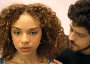 Evelyn com um olhar sério. Tomás aparece ao lado dela.