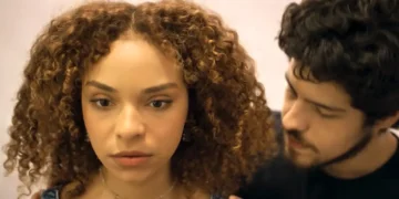 Evelyn com um olhar sério. Tomás aparece ao lado dela.