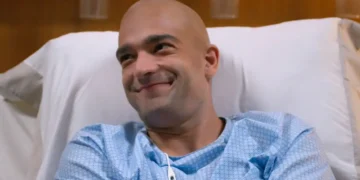 Afonso, personagem de Humberto Carrão, com o cabelo raspado, em uma cama de hospital, sorrindo.
