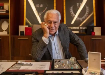 Arthur Brandão (Antonio Fagundes), da novela Quem Ama Cuida, aparece sorridente em seu escritório, sentado atrás de uma mesa com joias e pedras preciosas expostas, tendo ao fundo vitrines com colares luxuosos.